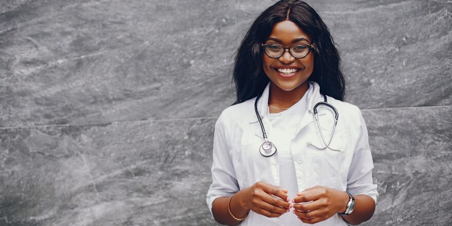 Stylish black doctor in a white uniform. Lady with stethoscope. Woman standing near gray wall